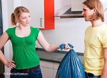 Eine Frau hält einem Mann in der Wohnung einen Müllsack hin, damit er ihn wegbringt