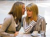 Zwei Frauen sitzen im Café: Die eine (links) flüstert der anderen etwas ins Ohr